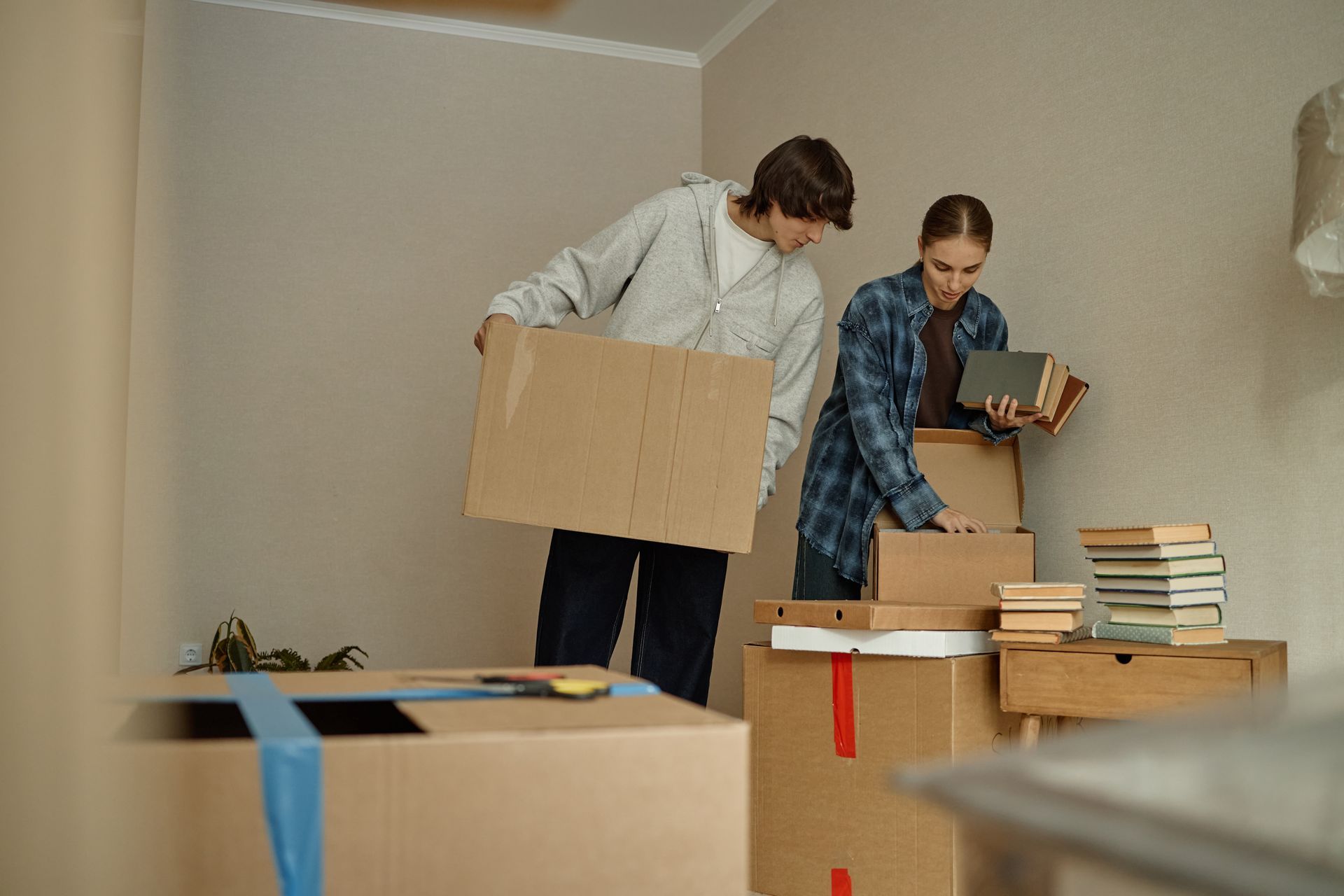 Two people packing boxes in a bare room, moving books and cardboard boxes.
