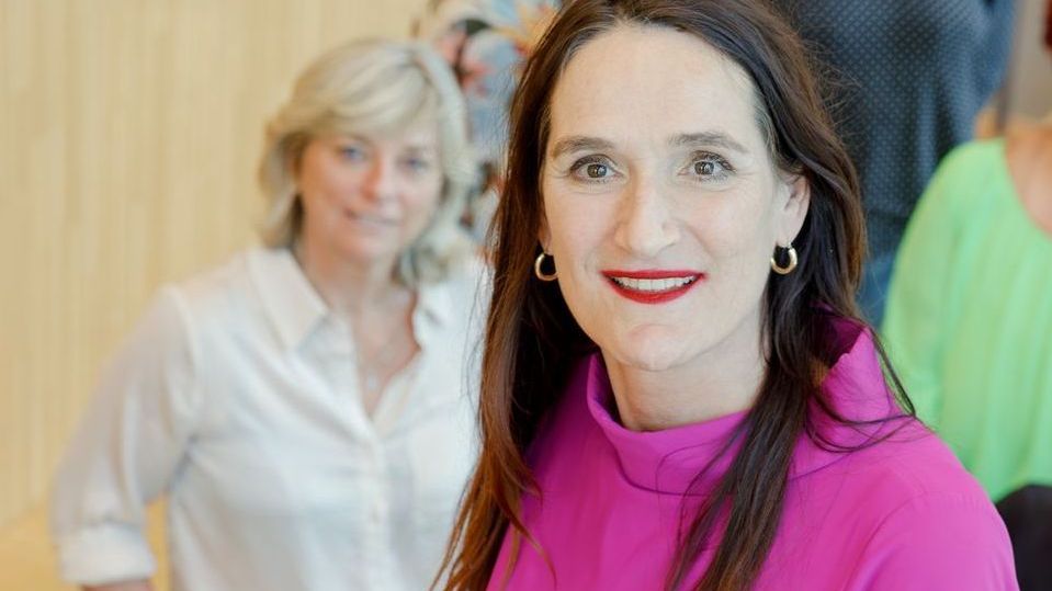Twee vrouwen in een kantoor, een van hen in een felroze top, lachend op de voorgrond.
