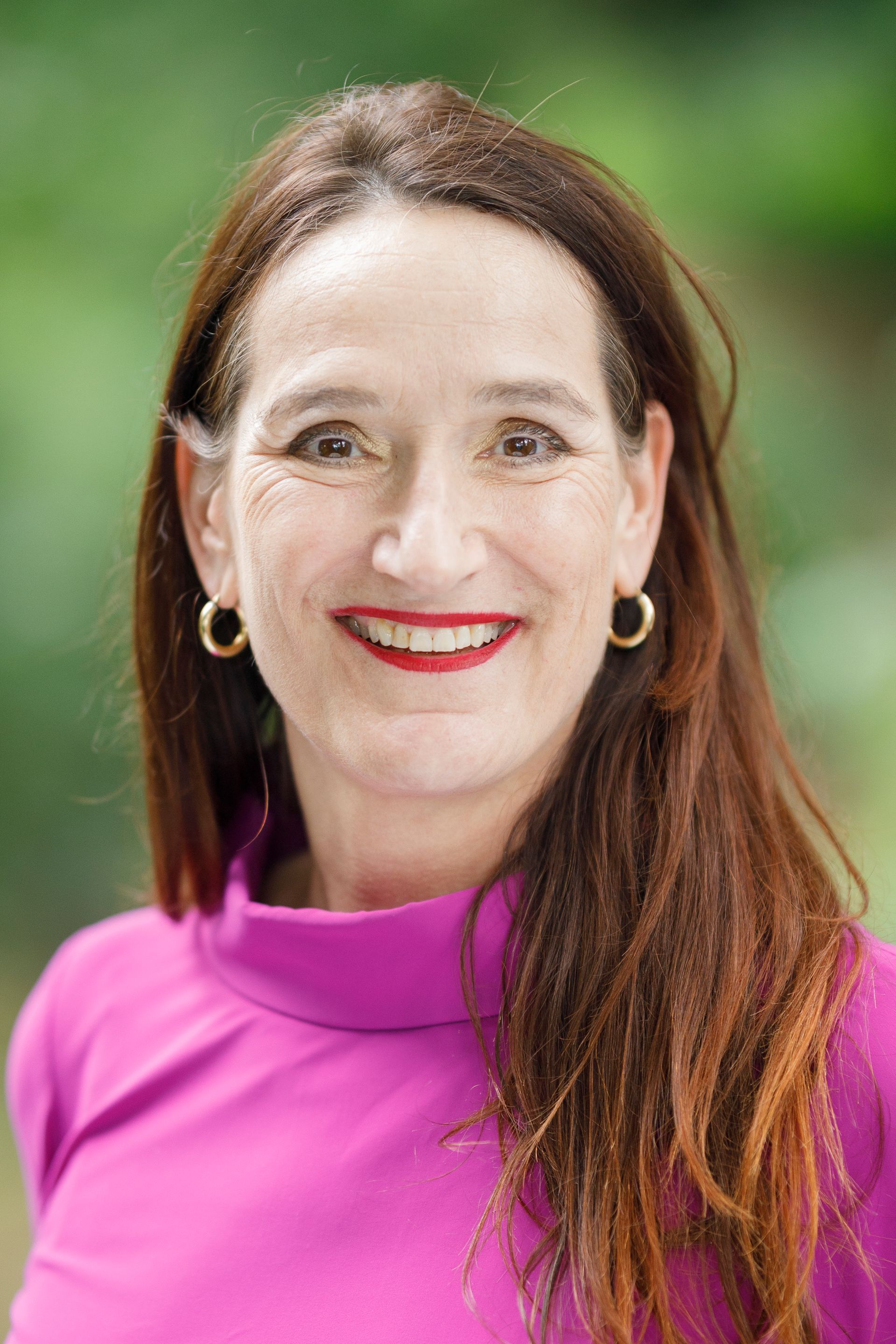Lachende vrouw in een magenta topje tegen een wazige groene buitenachtergrond.