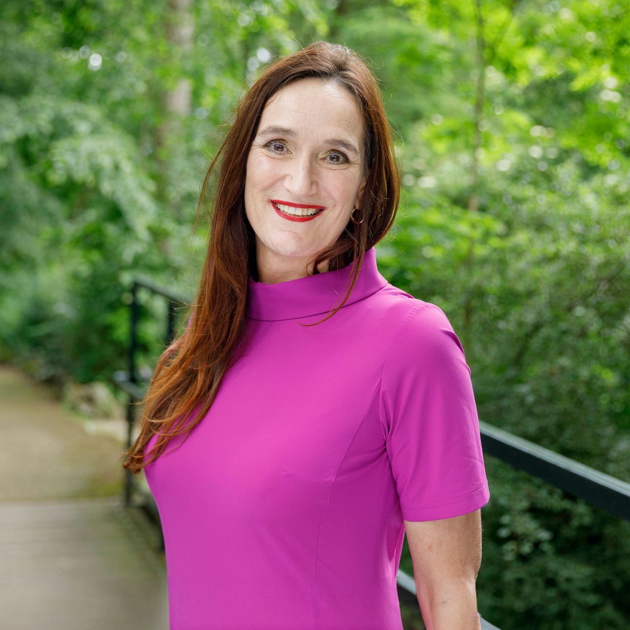 Vrouw in een felroze top lacht op een lommerrijk buitenpad.