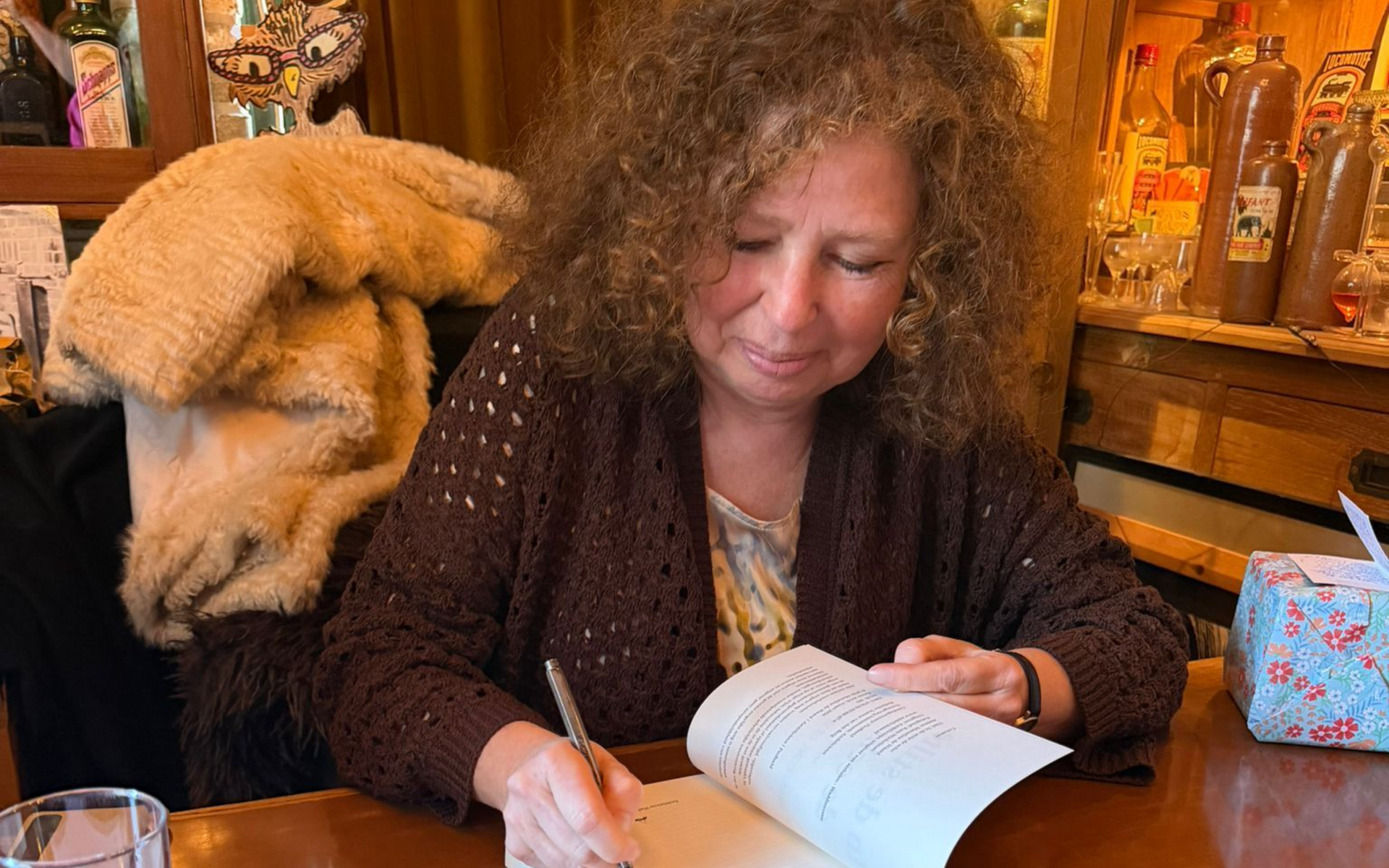 Een vrouw schrijft aan een houten tafel in een gezellige kamer met planken en een beige knuffeldier achter haar.