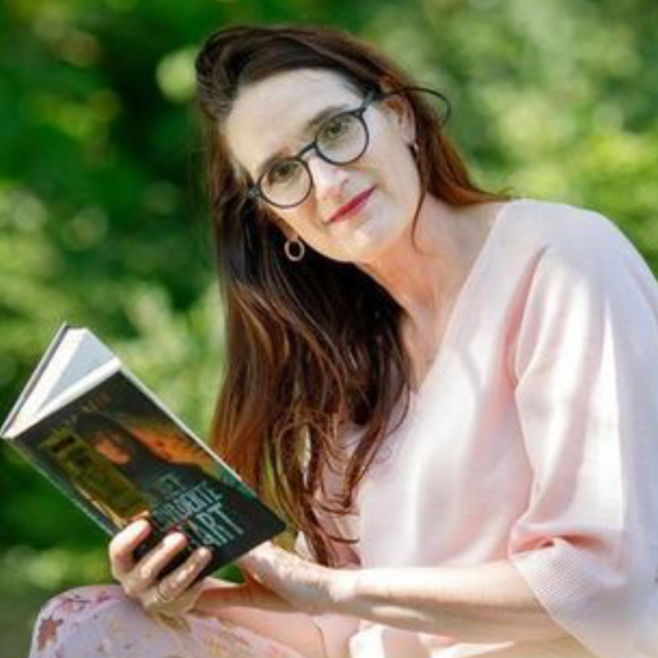 Dorine Holman met een licht roze truitje en een roze gebloemde rok terwijl ze in de camera kijkt en een boek in haar hand heeft. 