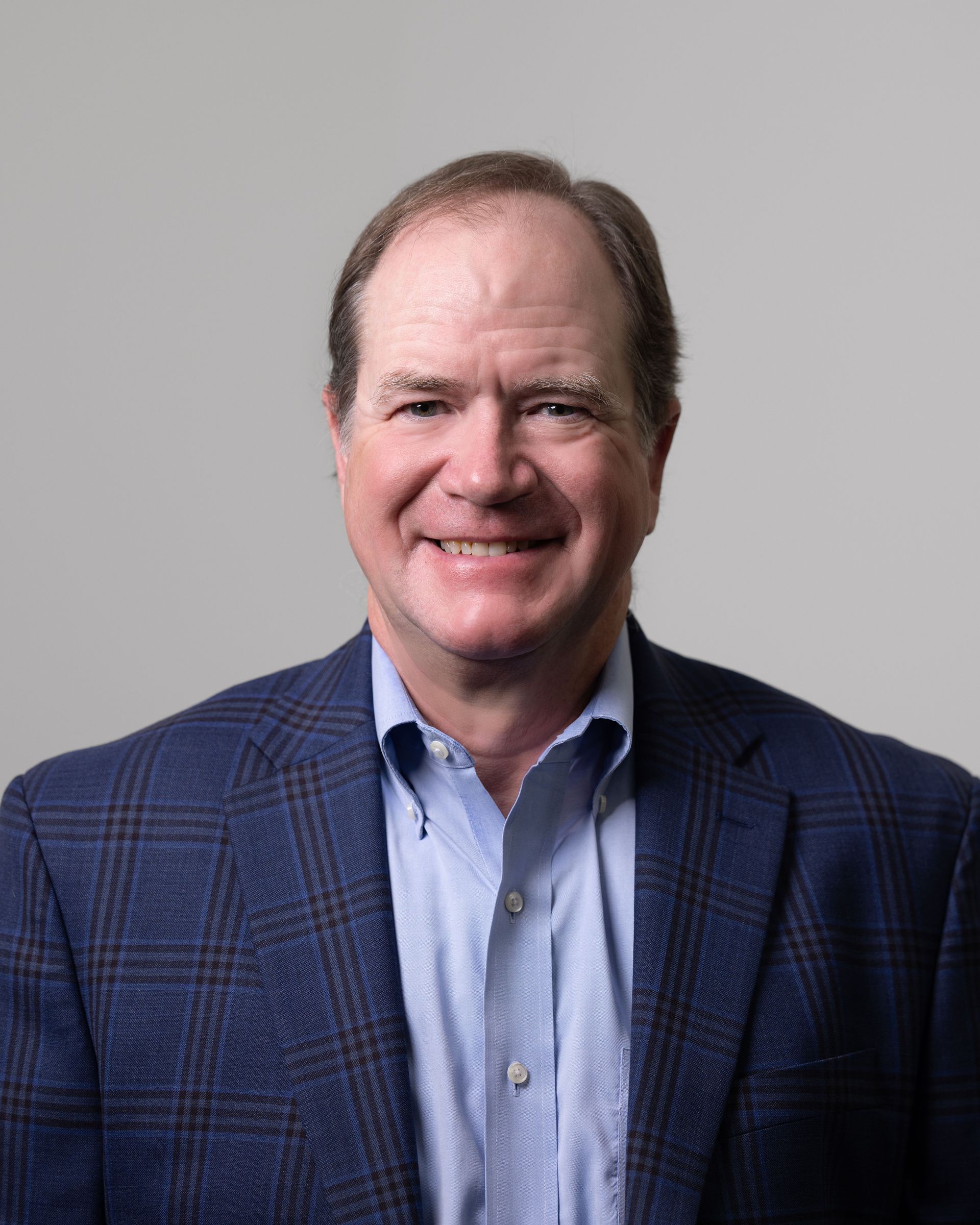 Smiling man in blue blazer and open-collared shirt, against a gray backdrop.