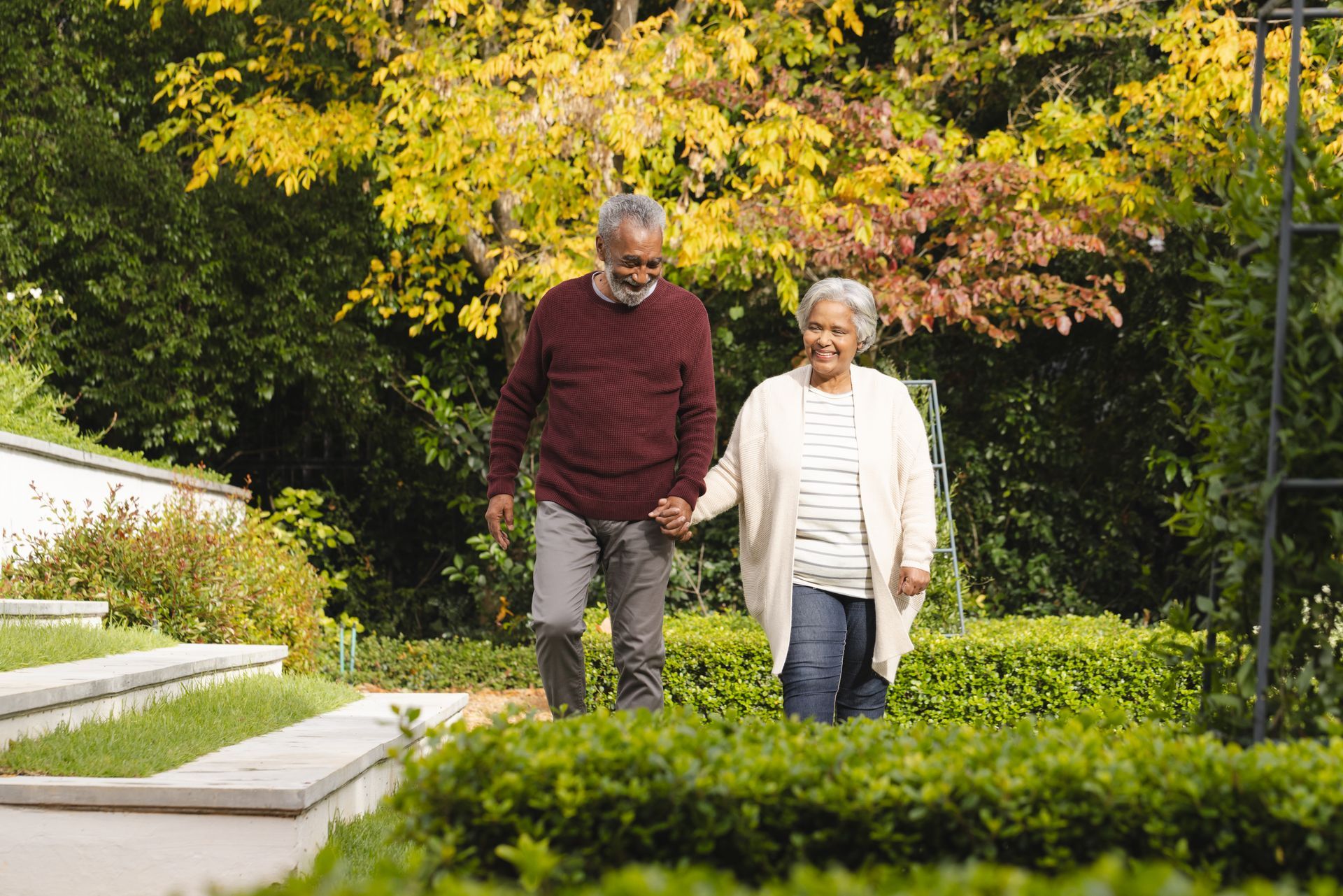 Couple walking hand-in-hand in a sunny garden. They are surrounded by green bushes, steps, and colorful autumn foliage.
