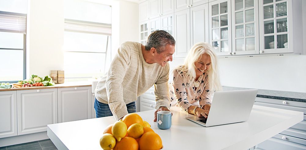 Couple smiles while looking at a laptop in a bright kitchen. A bowl of fruit sits nearby.