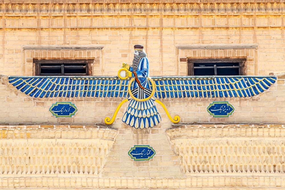 Faravahar symbol, winged figure with blue and white detail, on tan brick wall.