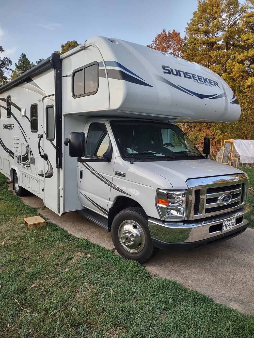 White Sunseeker RV parked on a grassy driveway. Fall foliage in the background.