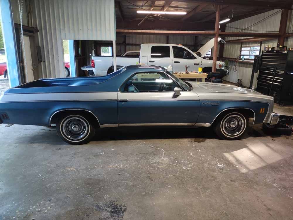 Blue and silver classic El Camino parked in a garage with a white pickup truck.
