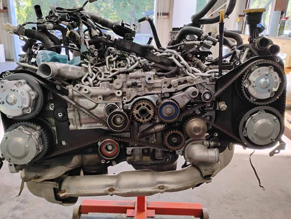 Subaru engine on a stand, showing belts, pulleys, and intricate wiring. Gray metal, black belts.