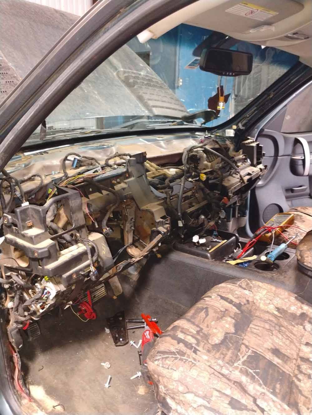 Dashboard of a vehicle disassembled, wires exposed, tools visible, camo seat.