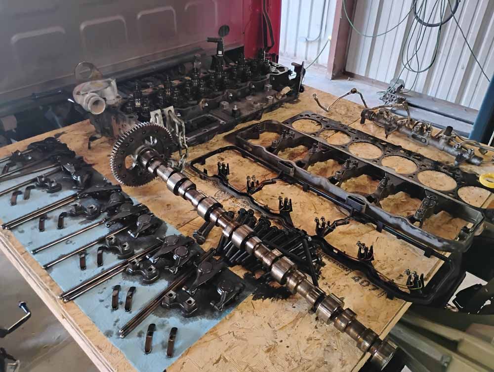 Disassembled engine parts laid out on a workbench. Various components, including the camshaft, are visible.