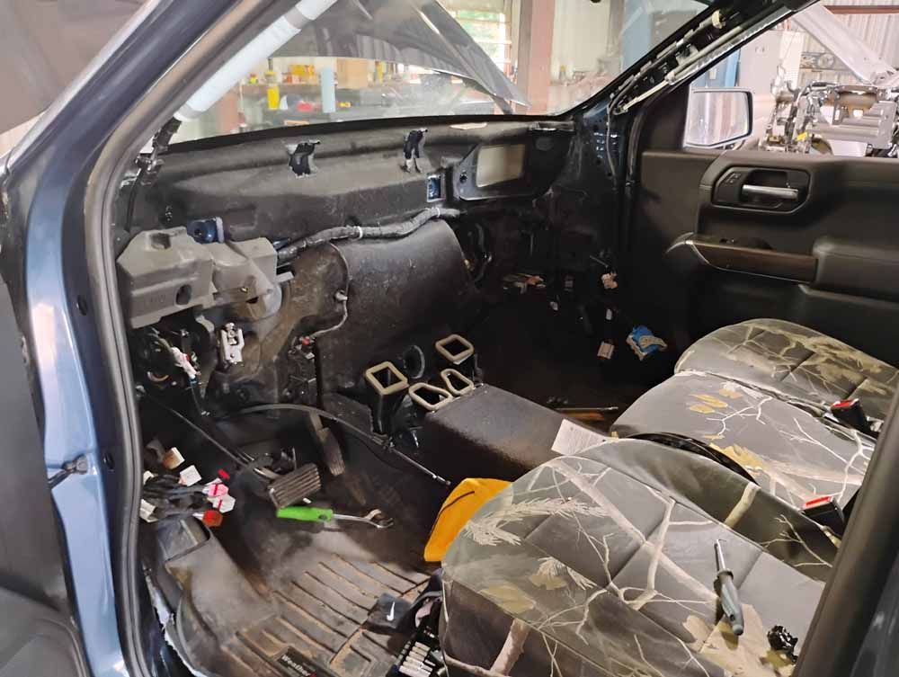 Interior of a disassembled vehicle, dashboard exposed. Black and gray components visible, camouflage seat covers. Tools and wires are present.