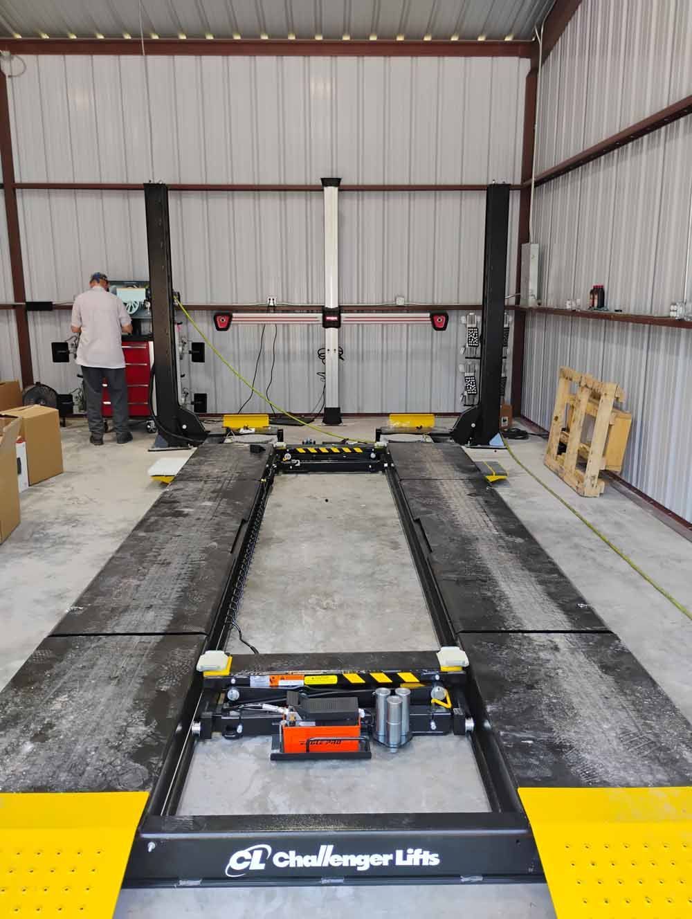 Car lift in a garage. A person works on the left. The lift is black, yellow, and silver.
