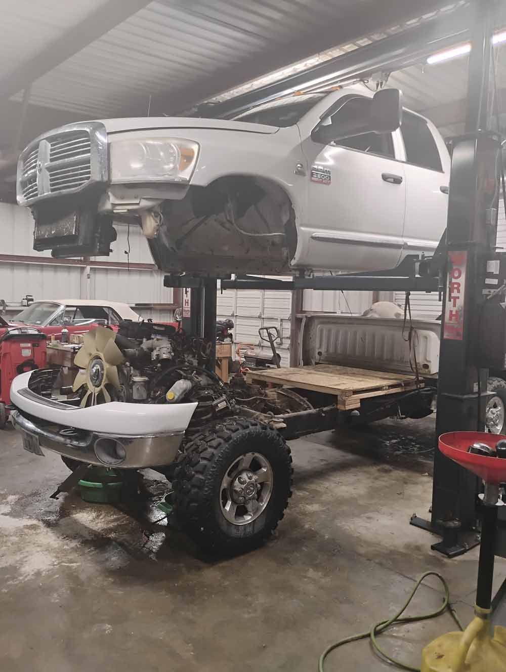 White pickup truck on a lift in a garage, undergoing repair. Front end and engine visible.