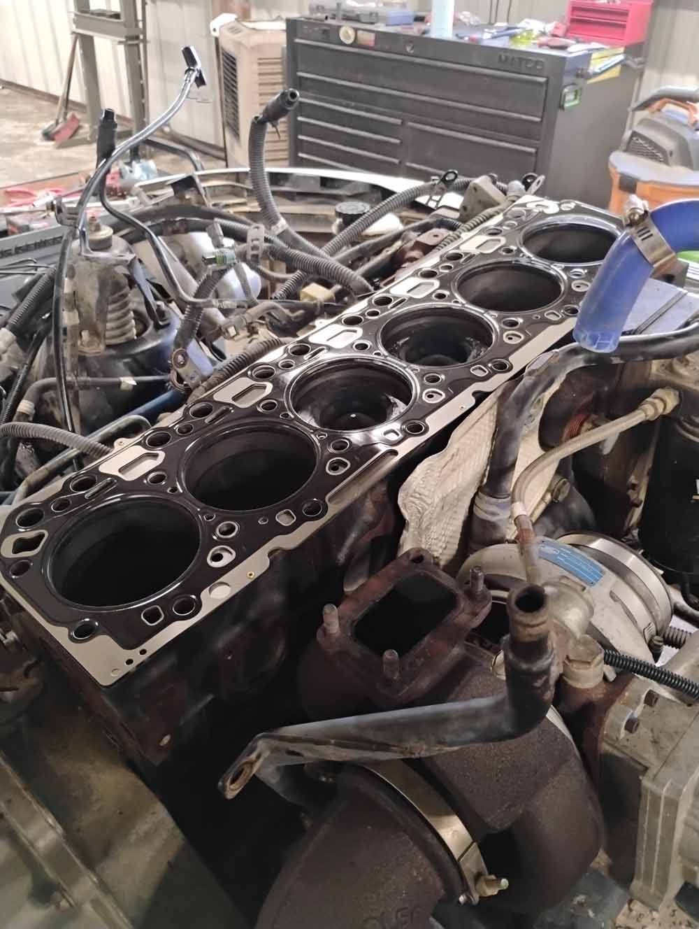 Engine block with open cylinders, various wires, hoses, and mechanical components visible in a garage setting.