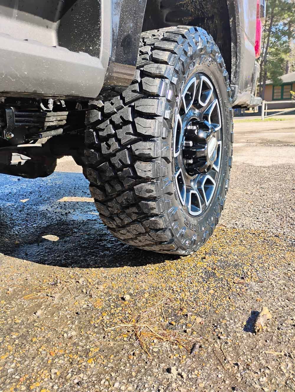 Close-up of a black truck's rear tire and wheel on a gravel surface. The tire has a deep tread.
