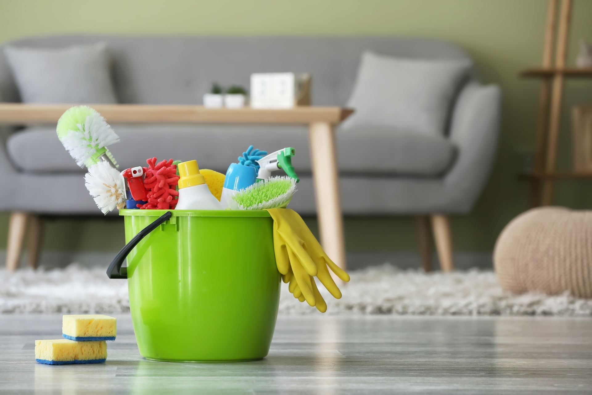 Green cleaning bucket with sponges and a brush on a living room floor, couch in background