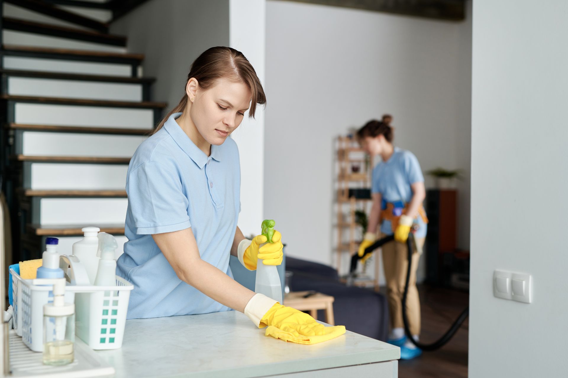 Two cleaners in a home: one wipes a table, the other vacuums in the background.