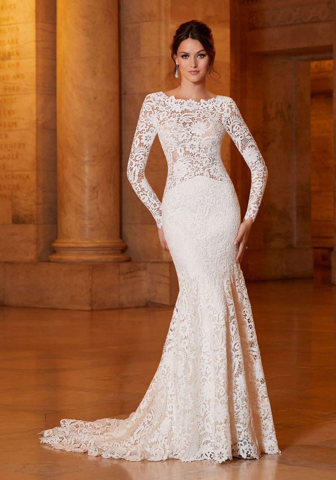 Una persona posa indossando un abito da sposa a sirena in pizzo bianco, lungo fino ai piedi e a maniche lunghe, all'interno di un edificio classico con colonne.