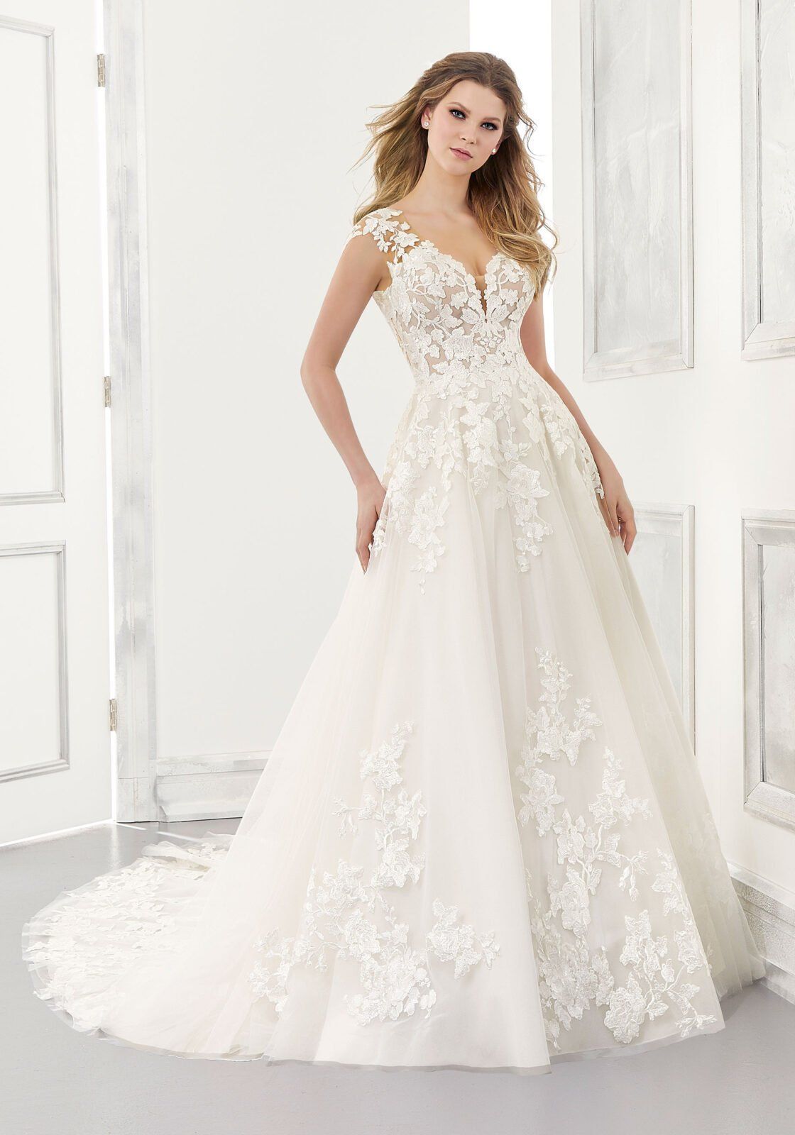 Una persona che indossa un abito da sposa bianco a trapezio con corpetto in pizzo, maniche corte e gonna fluida con dettagli in pizzo floreale.