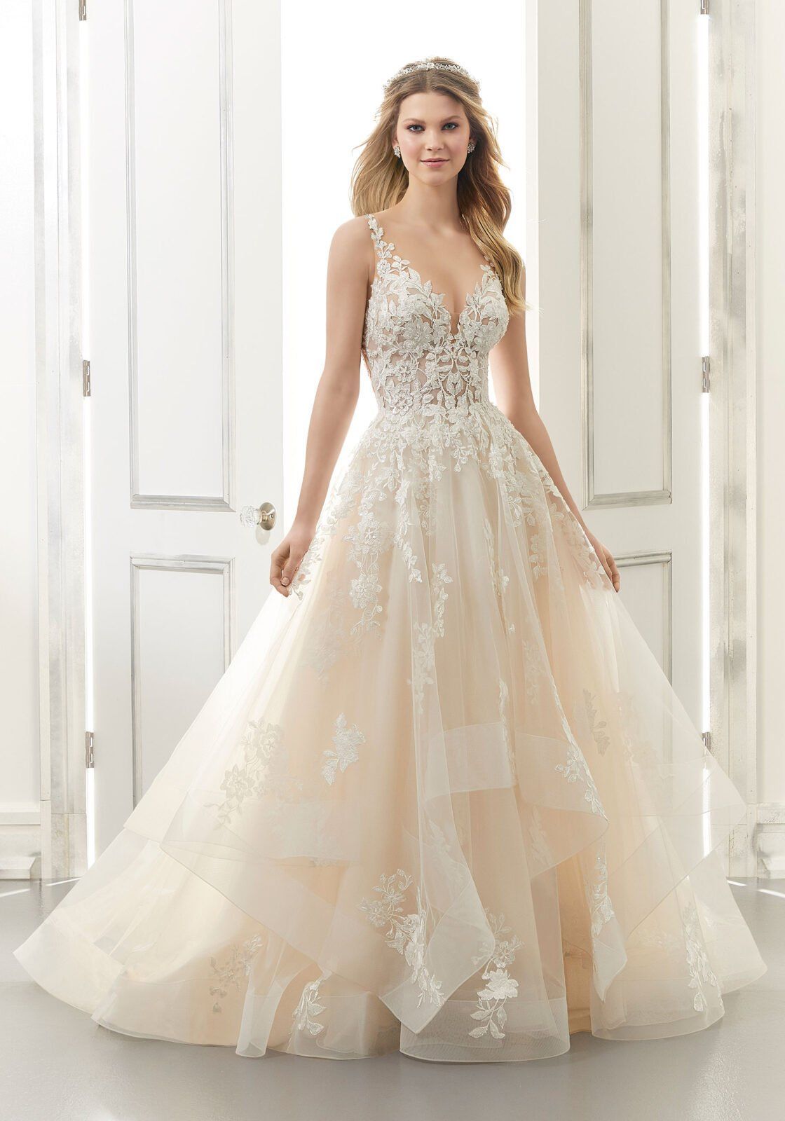 In una stanza luminosa, una persona indossa un abito da sposa beige a trapezio, lungo fino ai piedi, con corpetto in pizzo e gonna a strati di tulle.