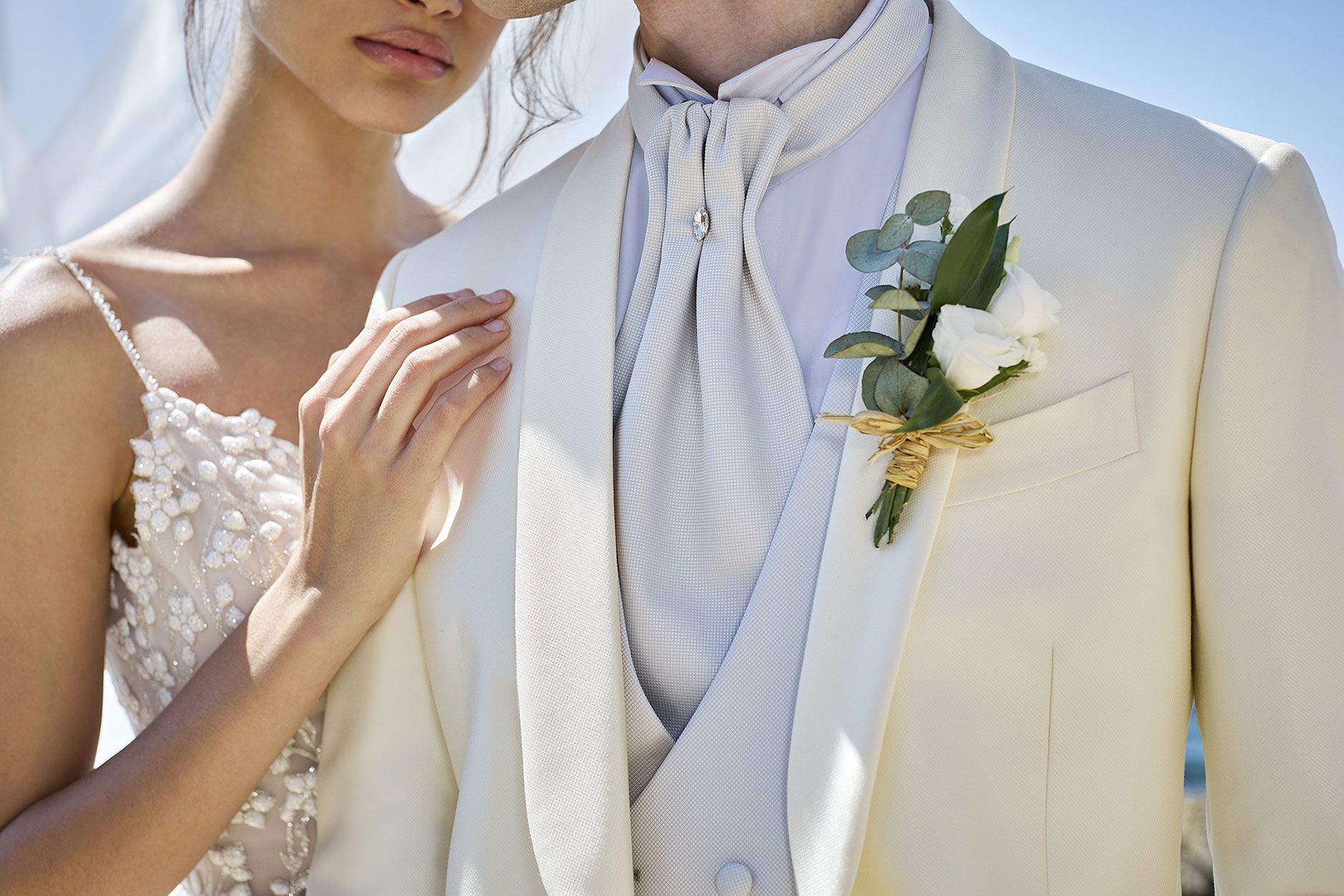 Una persona in abito da sposo color crema con una boutonnière floreale è in piedi accanto a una persona in abito da sposa di pizzo bianco.