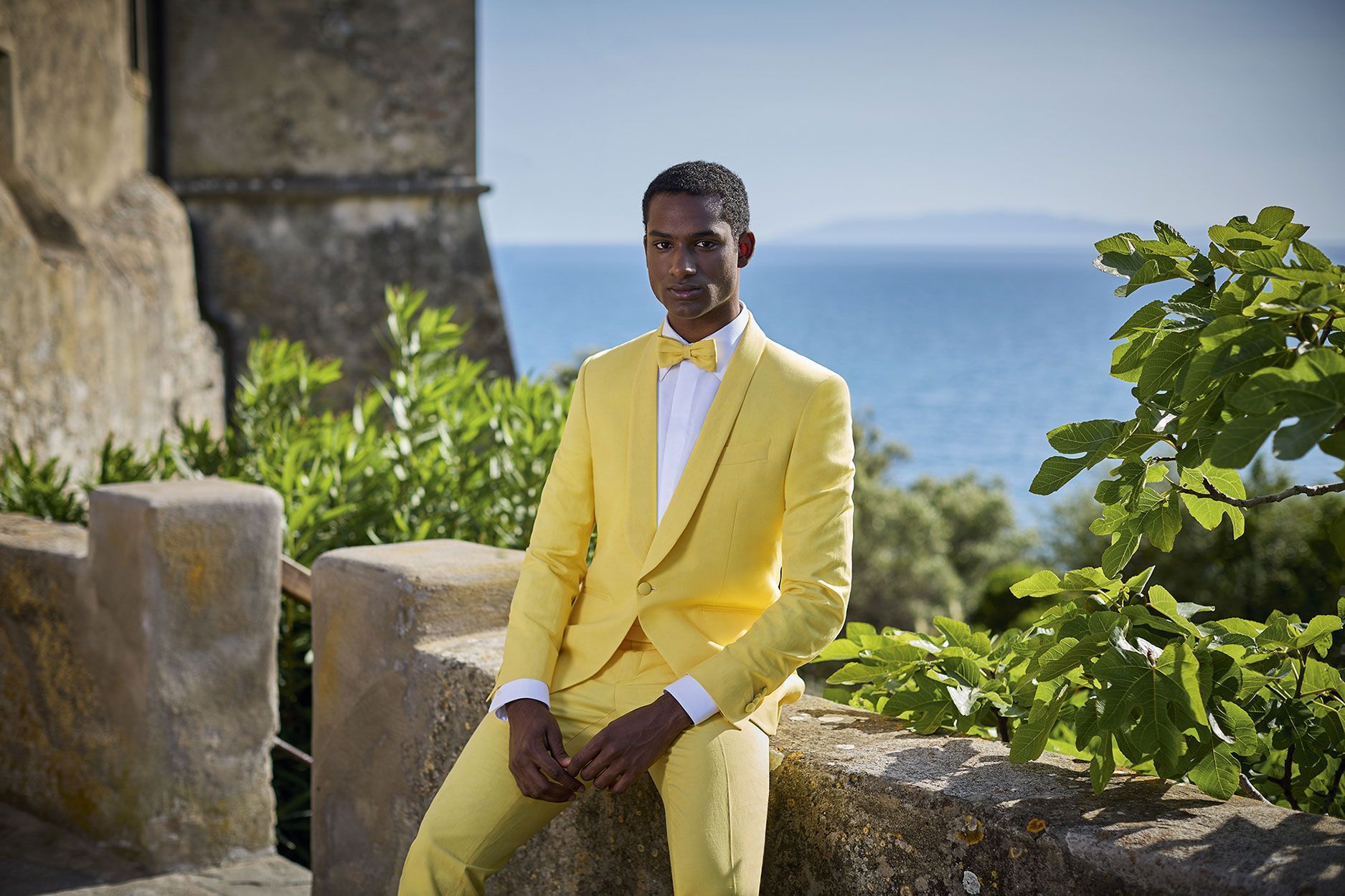 In una giornata di sole, una persona in abito giallo e papillon è seduta su un muretto di pietra con vista sul mare e sulla vegetazione lussureggiante.