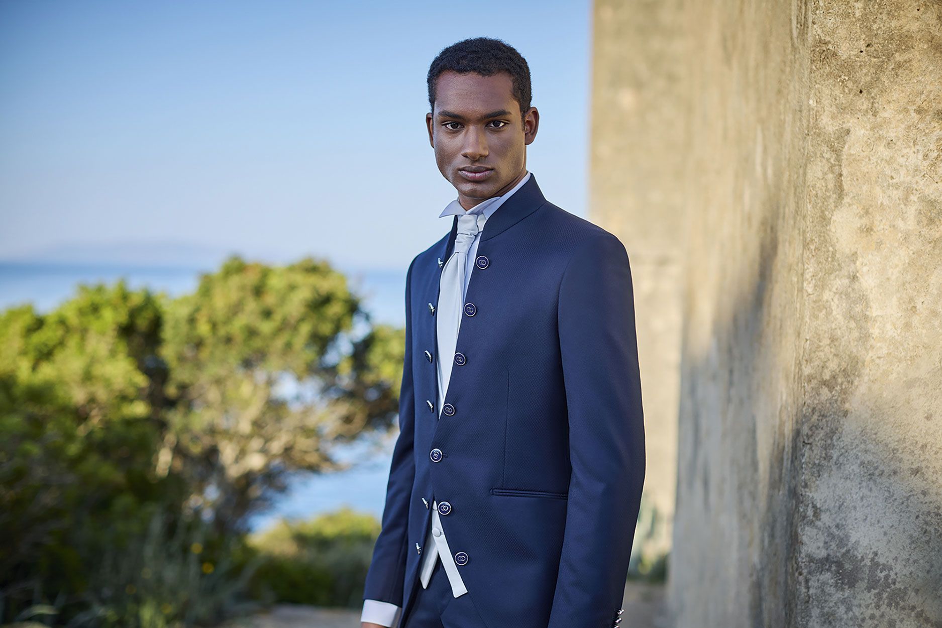 Una persona indossa una giacca blu scuro in stile Nehru sopra una camicia bianca, in piedi all'aperto contro un muro di pietra vicino al mare.