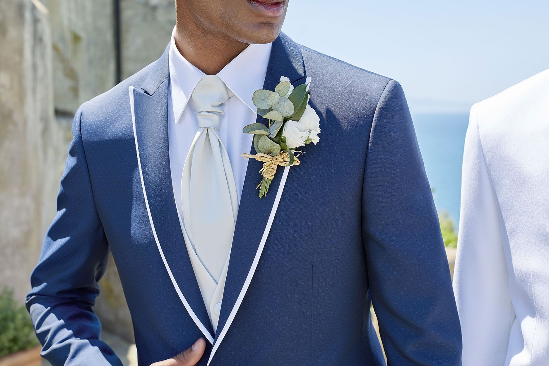 Una persona che indossa un abito blu testurizzato con profili bianchi, una camicia bianca, una cravatta chiara e un fiore all'occhiello.