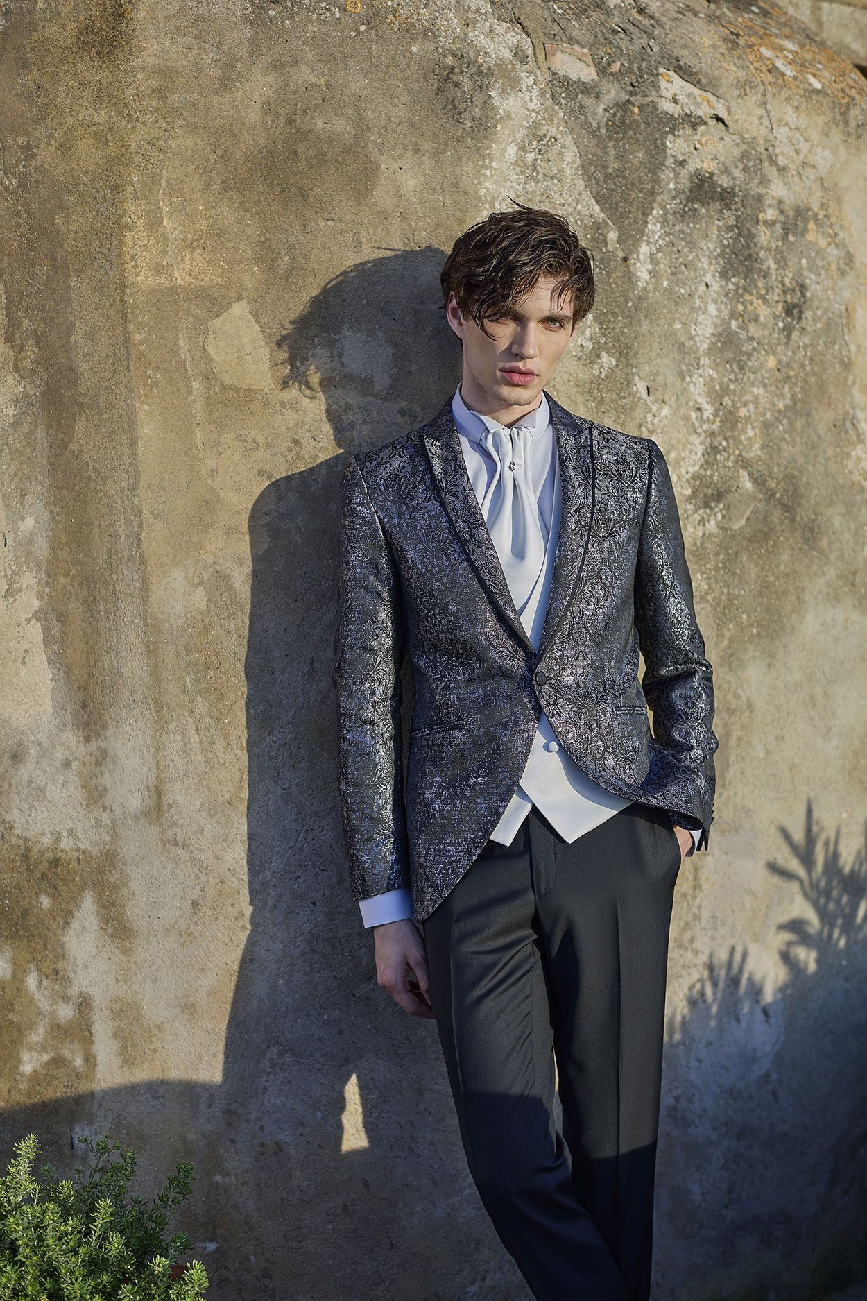 Una persona che indossa un blazer scuro con paillettes, una camicia chiara e pantaloni scuri è appoggiata a un muro di pietra ruvida sotto la luce del sole.
