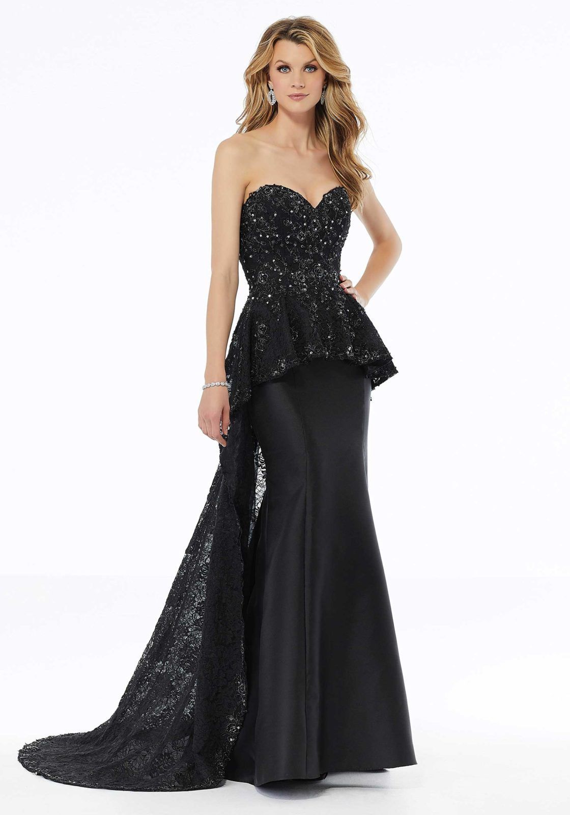 Una modella indossa un abito nero senza spalline, lungo fino ai piedi, caratterizzato da un corpetto peplum in pizzo tempestato di paillettes e da uno strascico in pizzo incorporato.