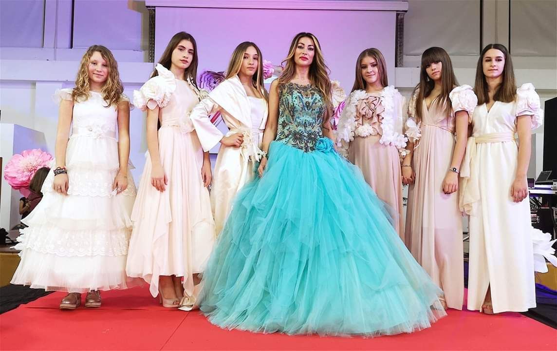 Sette persone in abiti da sera posano su una passerella rossa durante un evento al chiuso, con un grande abito turchese al centro.