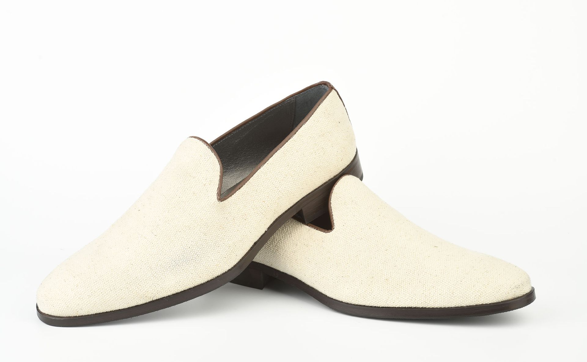 Un paio di scarpe slip-on color crema con texture e suola marrone scuro, ritratte su uno sfondo bianco uniforme.