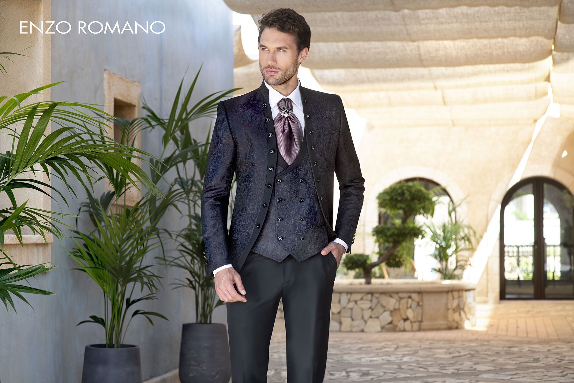 Un uomo in smoking blu scuro a fantasia, con gilet abbinato e cravatta a foulard color lavanda, in piedi in un cortile di pietra.