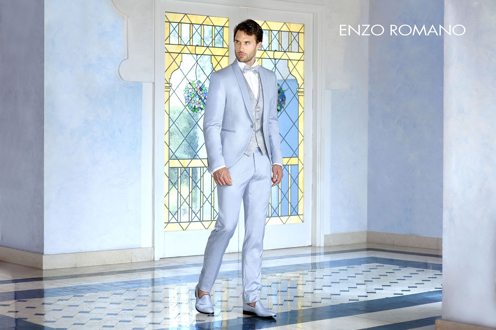 Un uomo che indossa un abito grigio chiaro con un gilet a fantasia si trova in una stanza piastrellata con una porta a vetri decorata.