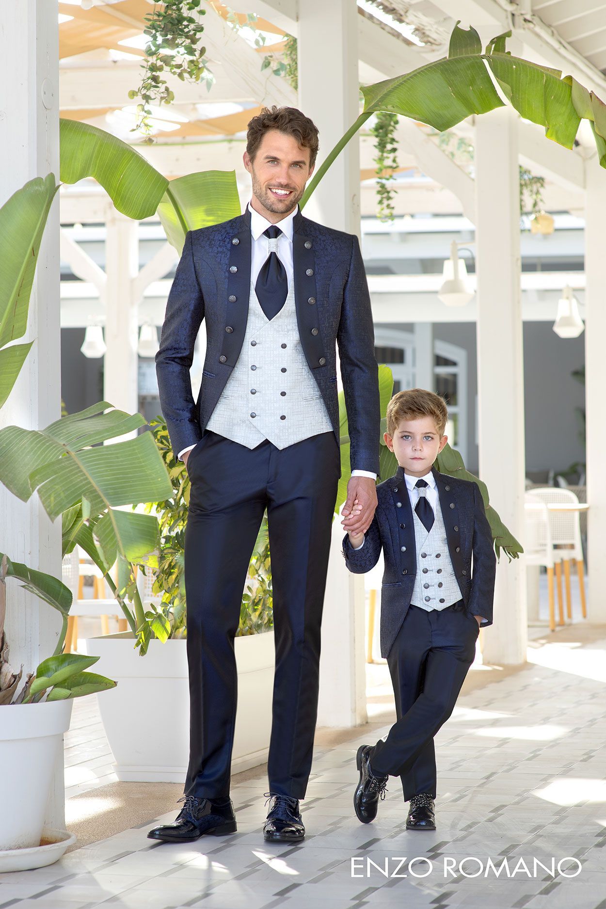 Un uomo e un bambino, vestiti con abiti blu scuro coordinati e gilet grigio chiaro, si tengono per mano su un luminoso viale all'aperto.
