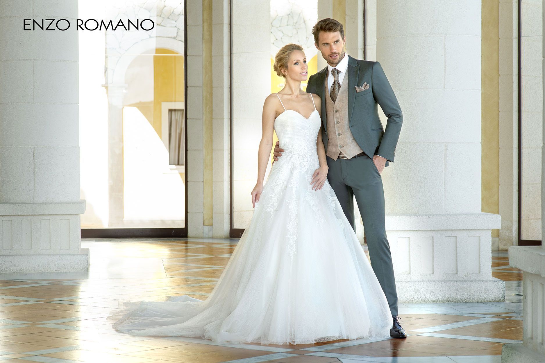 Una sposa in abito bianco e uno sposo in abito grigio sono in piedi insieme in un corridoio con colonne di pietra.