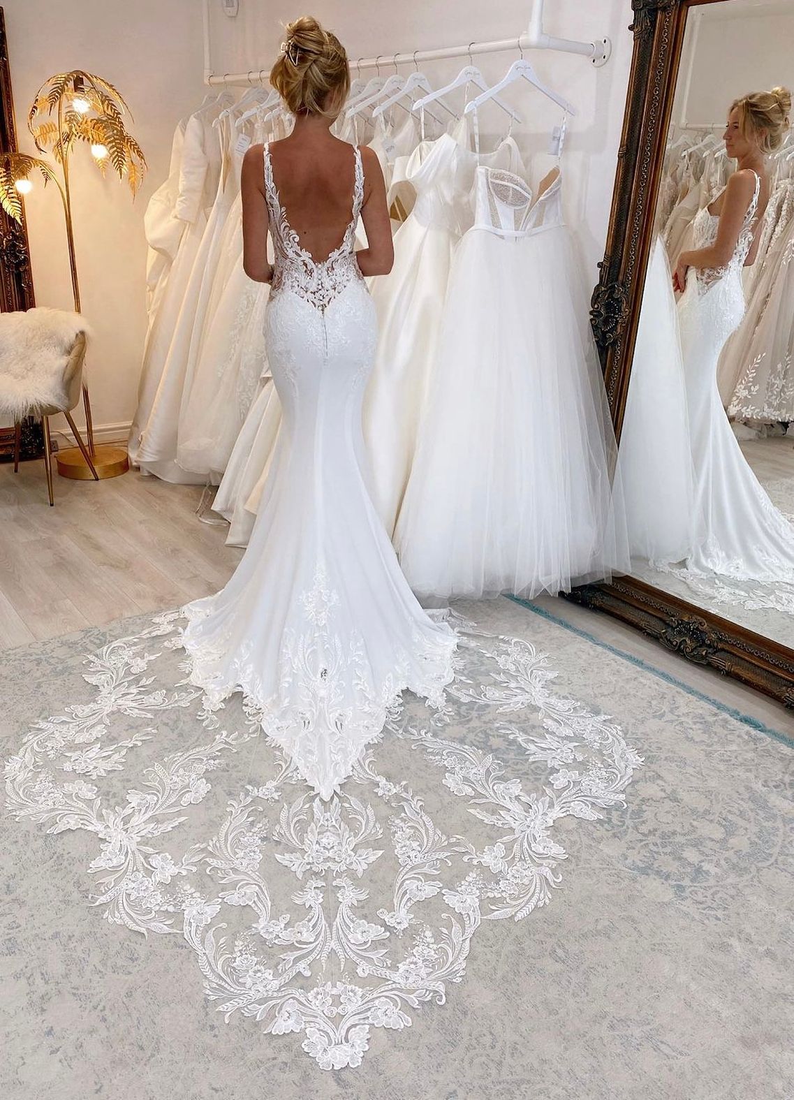 In un atelier da sposa, una persona indossa un abito da sposa bianco a sirena, senza schienale e con un elaborato strascico di pizzo.