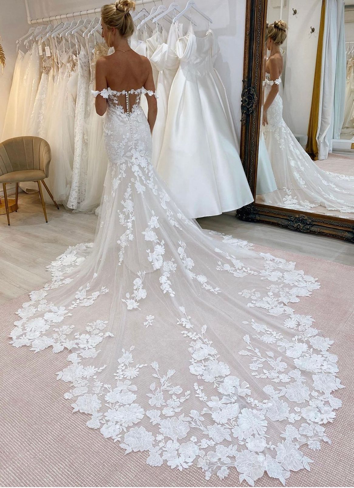 Una persona che indossa un abito da sposa bianco in pizzo, con spalle scoperte e un lungo strascico floreale elaborato, in un negozio di abiti da sposa.