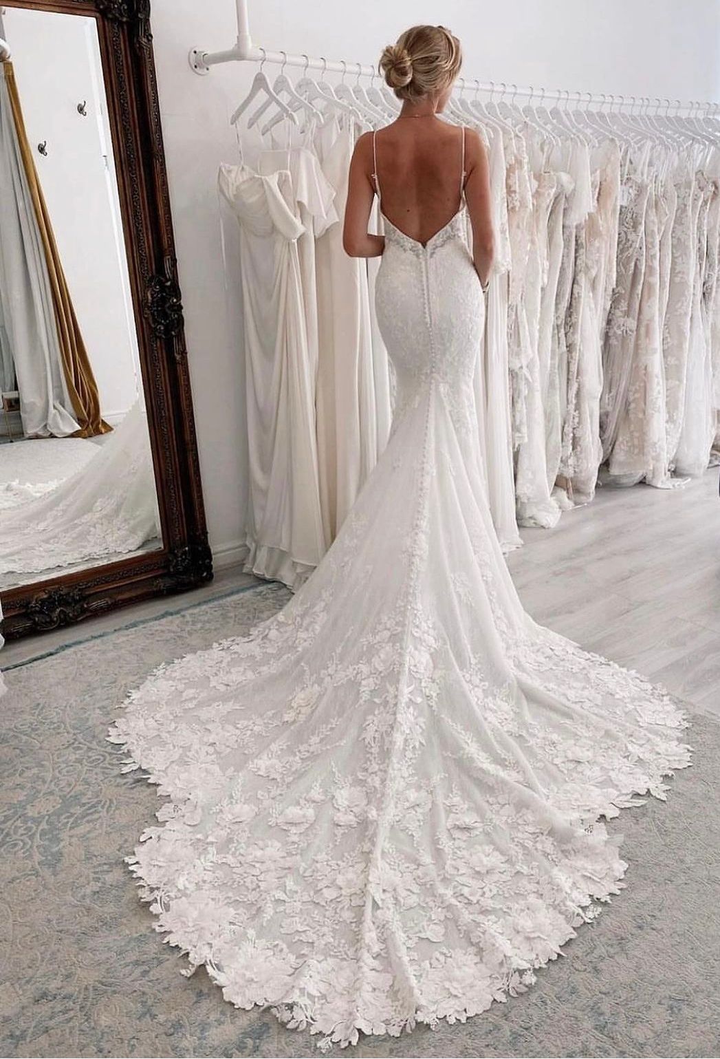 Una persona con indosso un abito da sposa a sirena, bianco, di pizzo, senza schienale e con un lungo strascico, si trova in una boutique di abiti da sposa.