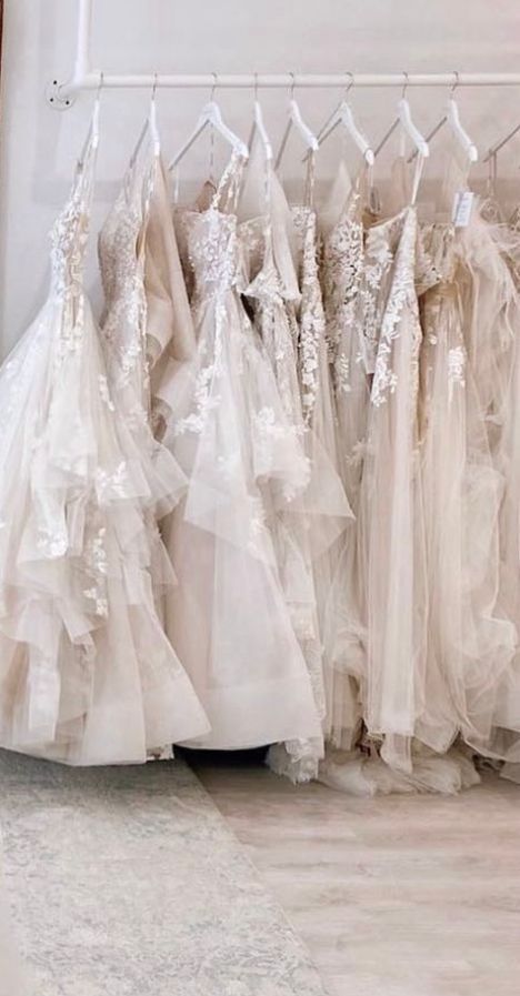 Una fila di eleganti abiti da sposa in pizzo, nelle tonalità del bianco e del crema, appesi a un appendiabiti bianco contro una parete neutra.