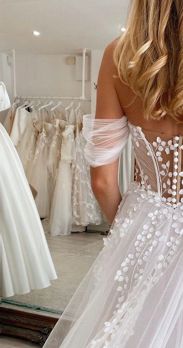 Una persona si trova in un negozio di abiti da sposa, indossando un abito da sposa bianco, con le spalle scoperte e decorazioni floreali.