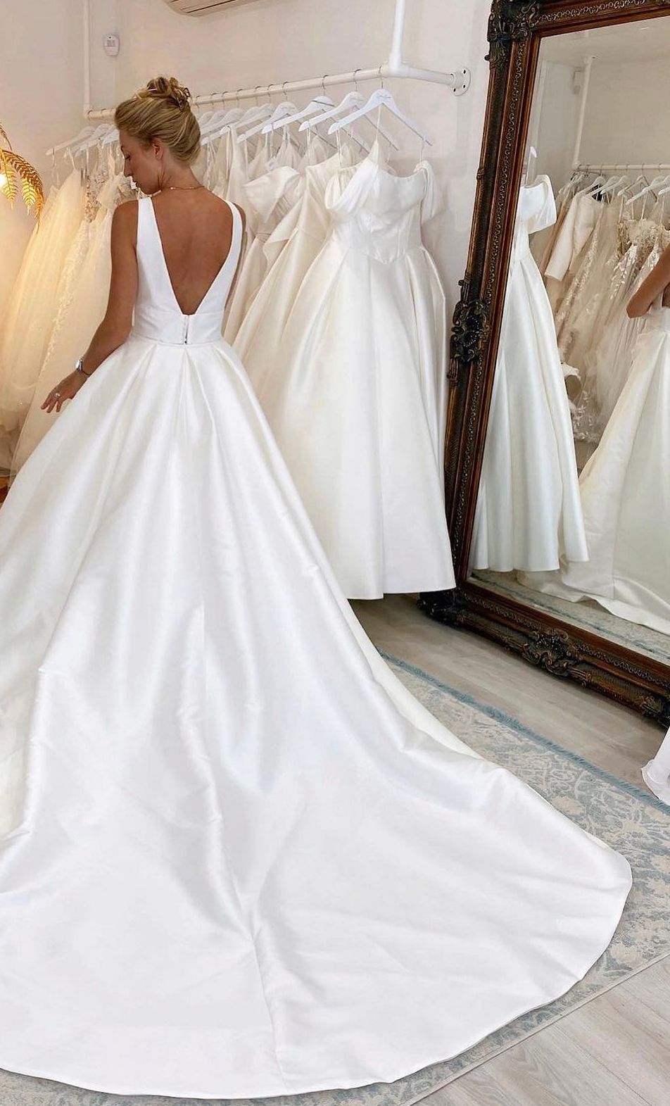 Una persona che indossa un abito da sposa bianco in raso con scollo a V sulla schiena e lungo strascico, in piedi in una boutique di abiti da sposa davanti agli specchi.