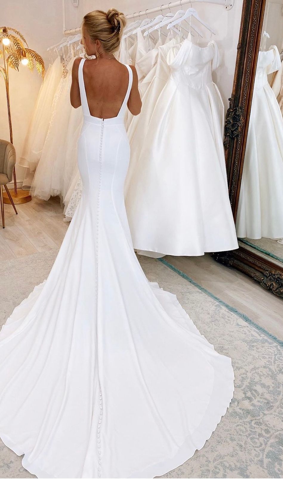 Una persona che indossa un abito da sposa bianco, lungo fino ai piedi e senza schienale, con un lungo strascico, in piedi in un negozio di abiti da sposa.