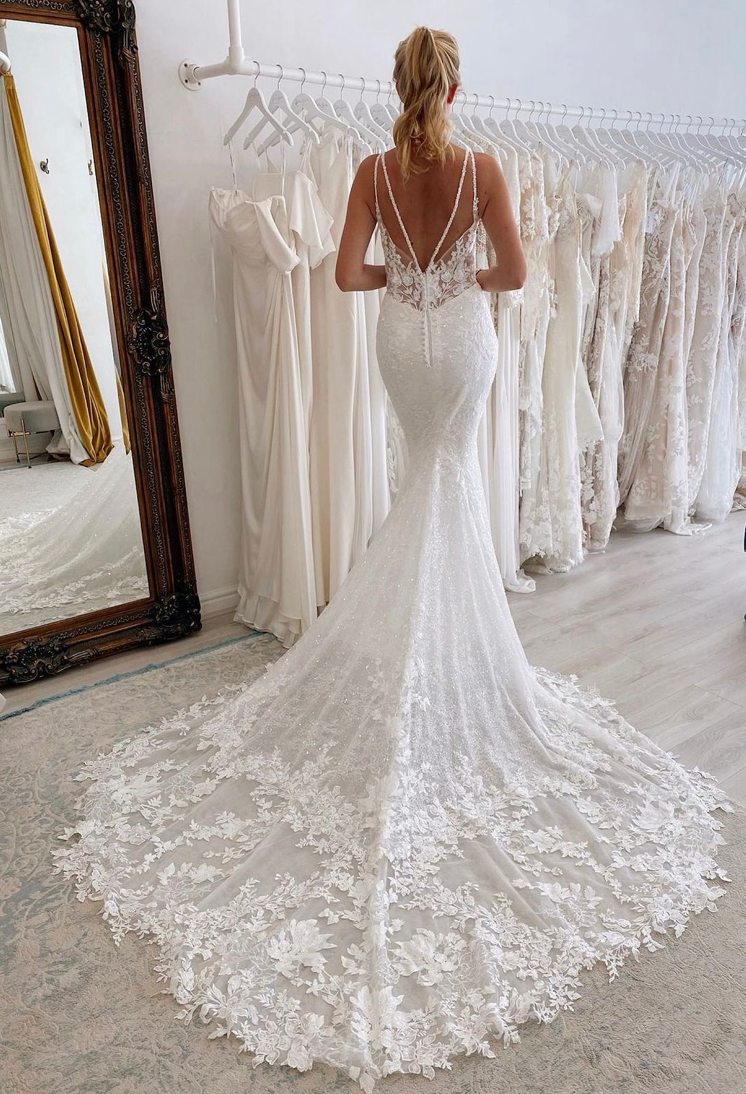 Una persona indossa un abito da sposa aderente in pizzo con un lungo strascico, in piedi in una boutique da sposa con specchi.