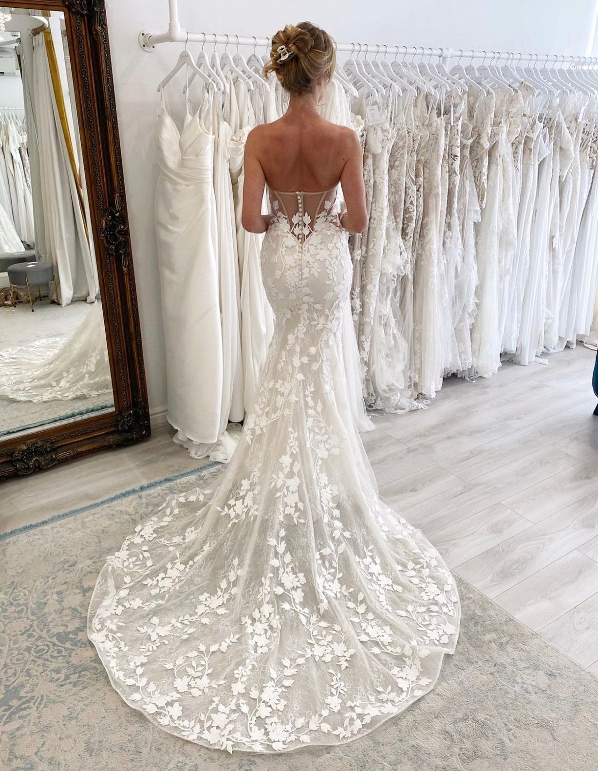 Una persona con indosso un abito da sposa aderente in pizzo con un lungo strascico si trova in un negozio di abiti da sposa, circondata da vestiti appesi ad appendiabiti.