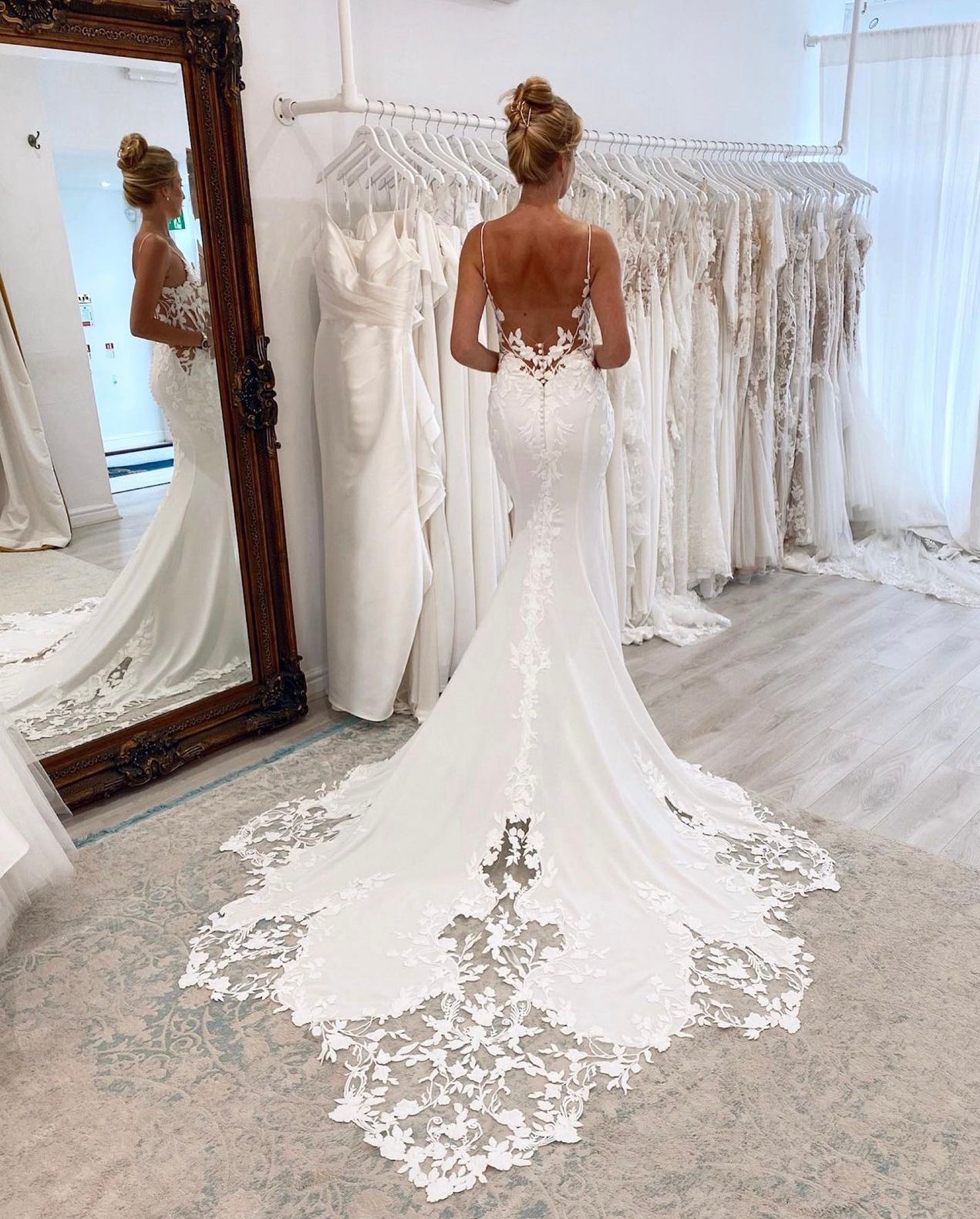 Una persona si trova in un negozio di abiti da sposa, indossando un abito da sposa bianco, senza schienale, a sirena, con un lungo strascico rifinito in pizzo.