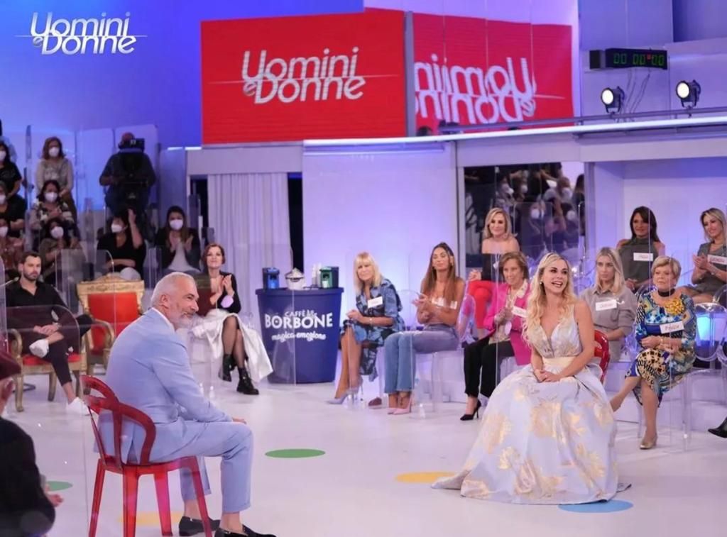 Lo studio televisivo allestito per il programma Uomini e Donne, con un partecipante seduto e alcuni membri del pubblico sullo sfondo.