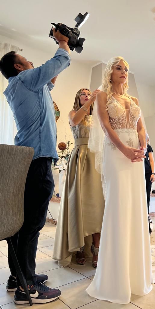 Una sposa in abito bianco e velo rimane immobile mentre un fotografo la filma e un'assistente le sistema il velo.