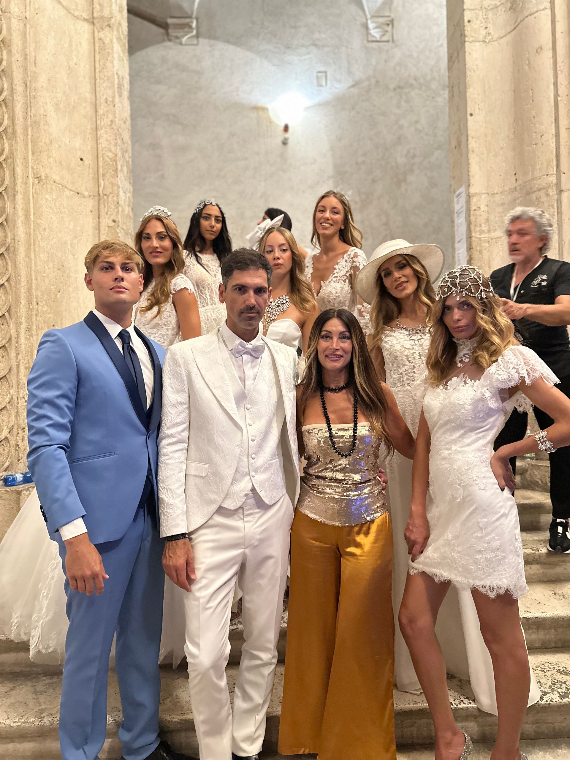 Un gruppo di persone posa su una scalinata di pietra: un uomo in abito blu, un uomo in abito bianco e diverse persone in eleganti abiti bianchi e dorati.